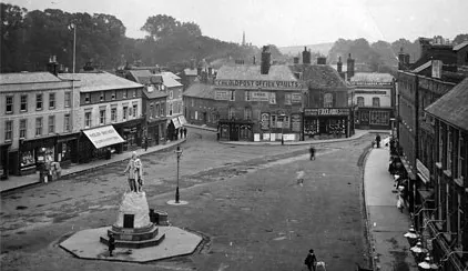 Old photograph looking down on a town square with a statue, shopfronts and a few people walking